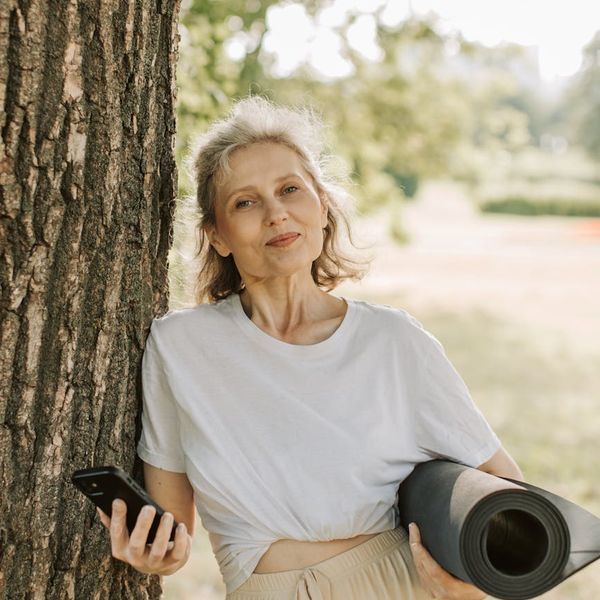 Person of diverse background smiling and holding a yoga mat outdoors.